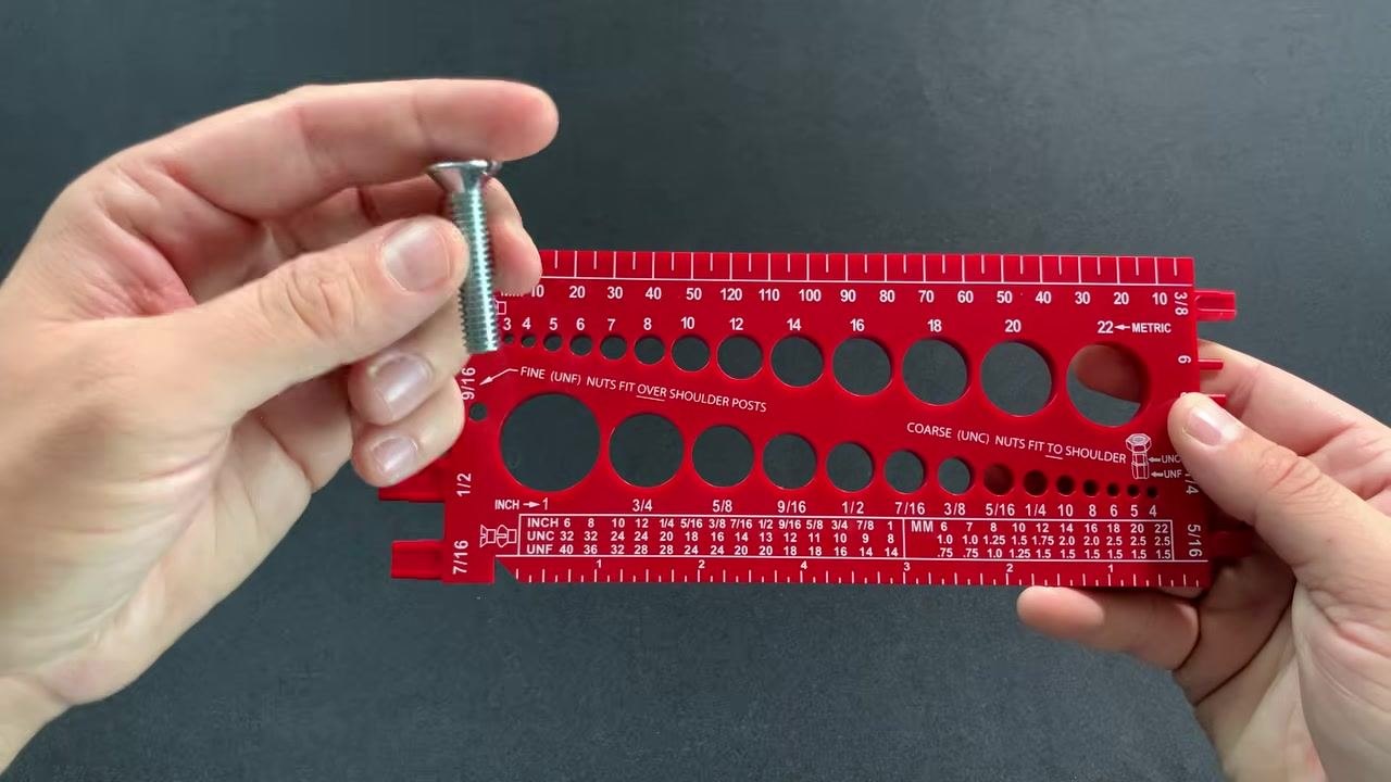 A hand holds a slotted flathead machine screw above the red gauge, preparing to insert it into the diameter holes, with a nut visible on the table.