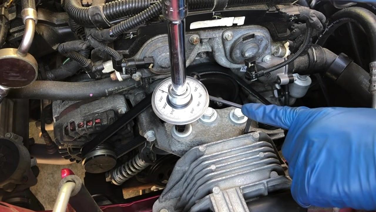 Gloved hand holding the torque angle gauge and socket, positioning it onto a bolt in a car engine bay with various engine components and wiring visible