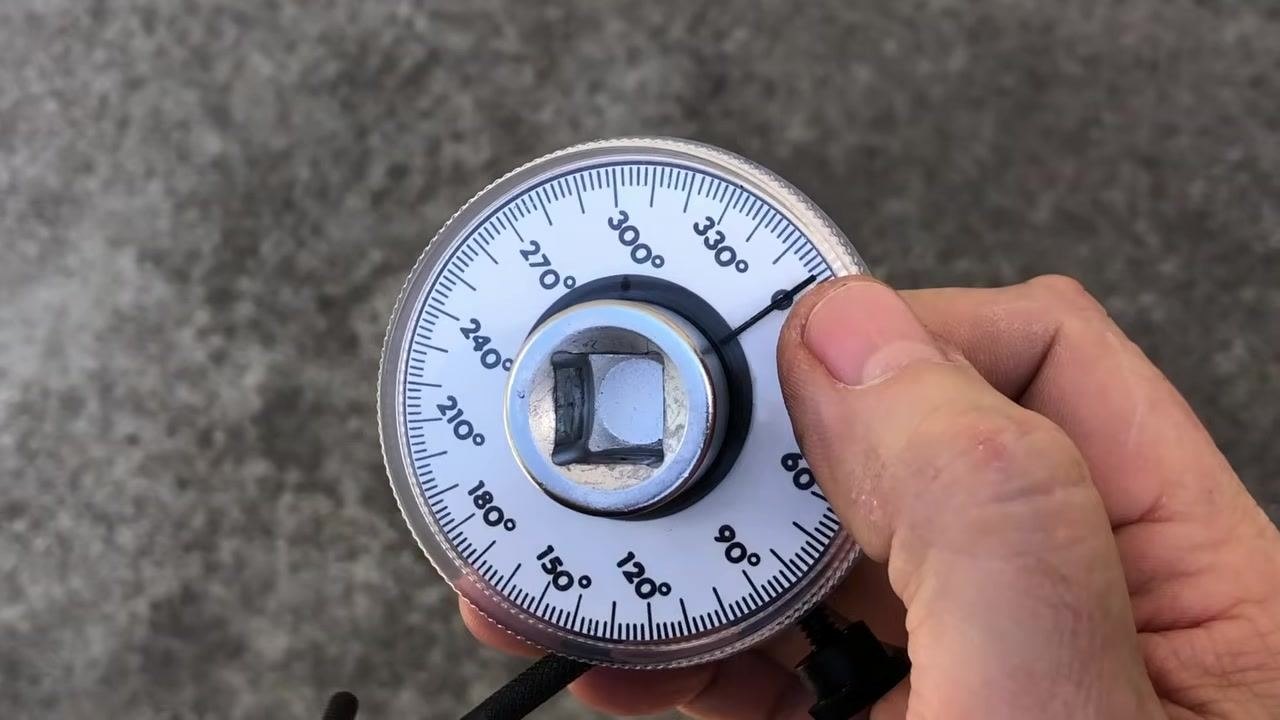 Close-up of a hand holding the OTC 4554 torque angle gauge, showing the degree markings (0° to 360°), central square drive, and pointer against a concrete background