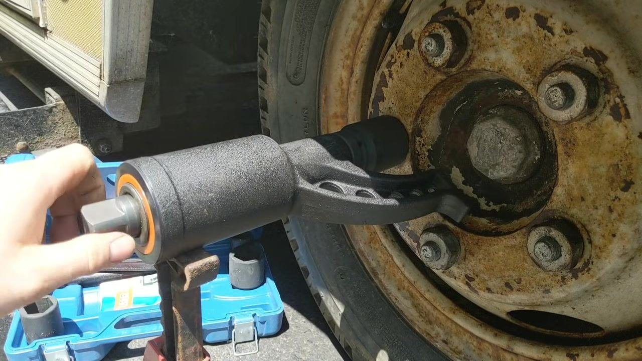 Torque multiplier socket attached to a rusty lug nut on a large vehicle wheel, blue tool case open on the ground, jack stand supporting the tool, and additional sockets visible