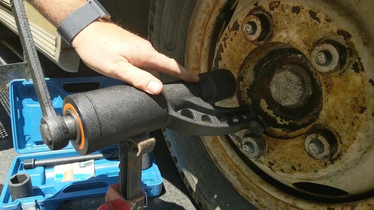 Hand holding the torque multiplier with the nut visibly loosened, blue case and jack stand below