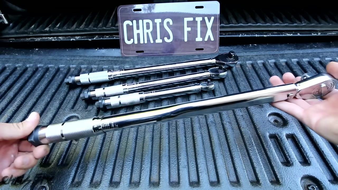 A torque wrench is held in both hands above a set of other torque wrenches and a breaker bar on a truck bed. "CHRIS FIX" license plate in the background.