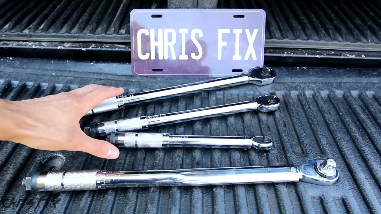 A hand holds a socket and extension, while another hand holds two black extension bars. Several torque wrenches and a breaker bar are laid out on a truck bed. "CHRIS FIX" license plate in the background.