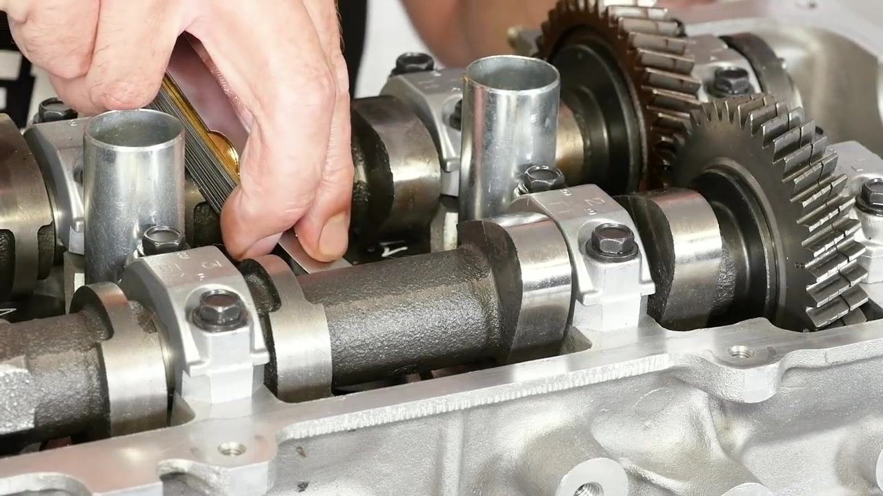 A hand holds a set of feeler gauges while inserting one between camshaft components in an engine. The camshaft, gears, and bolts are visible.