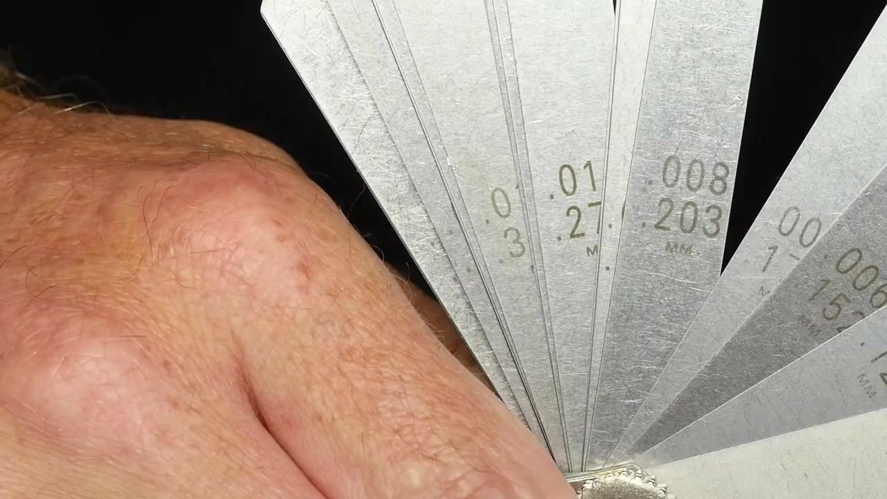 A close-up of several feeler gauges fanned out, showing thickness markings in both metric and imperial units. A hand holds the set.