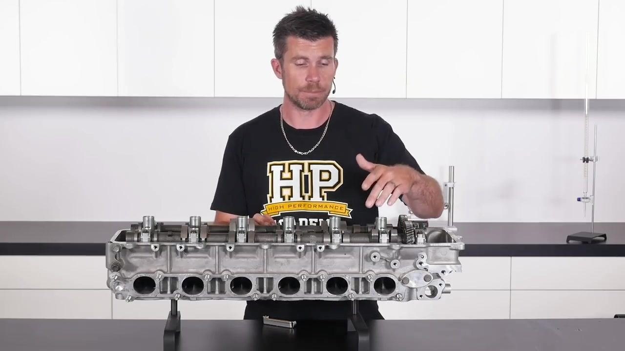 The instructor stands behind a Toyota 1FZ cylinder head, pointing to the area where the exhaust valve clearance will be measured. The cylinder head is mounted on a bench.