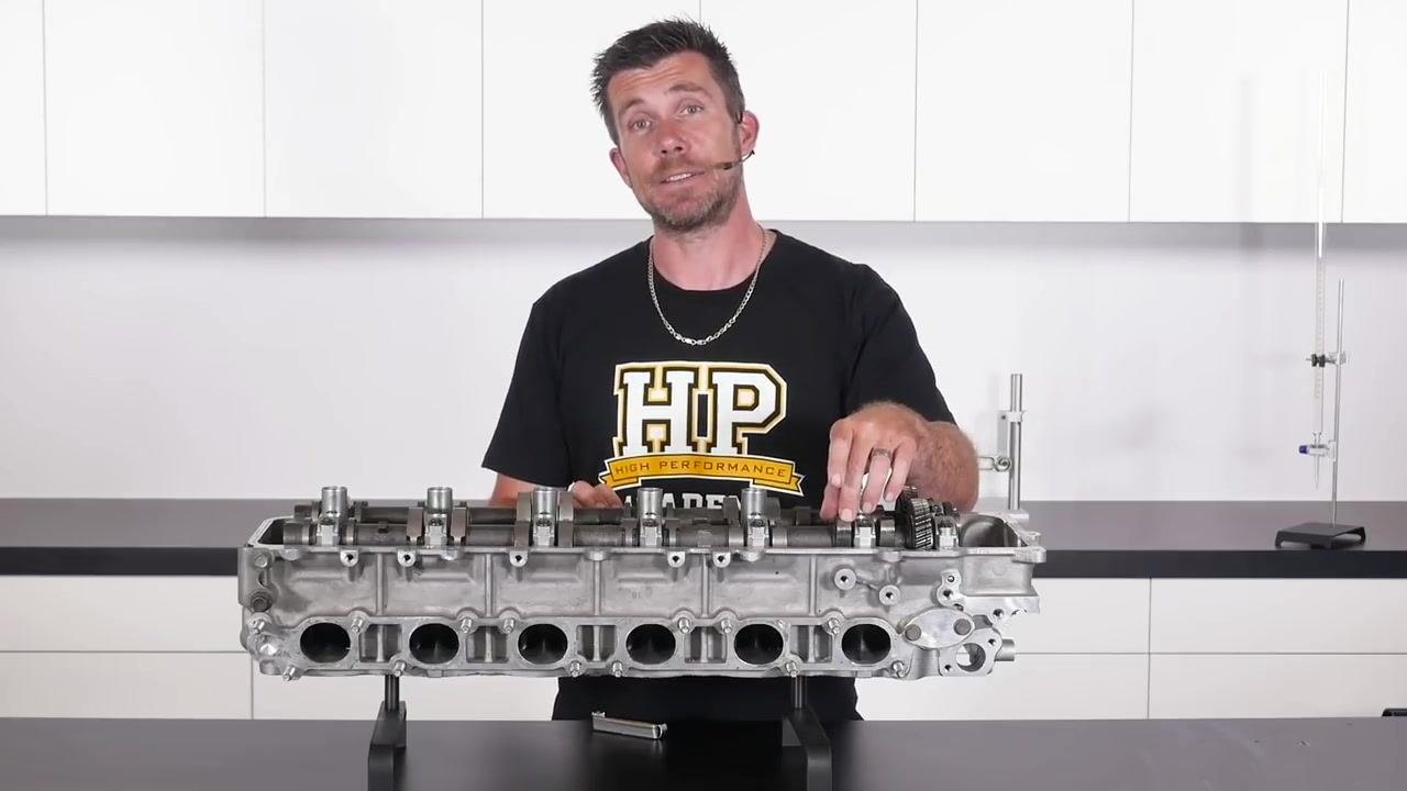 The instructor points to the camshaft lobe and bucket area on the cylinder head, preparing to demonstrate where the feeler gauge will be inserted.