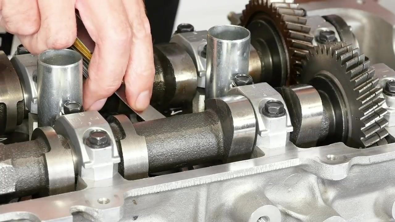 A close-up shows a hand inserting a feeler gauge between the camshaft lobe and the bucket on the cylinder head. The camshaft, cam caps, gears, and part of the feeler gauge set are visible.