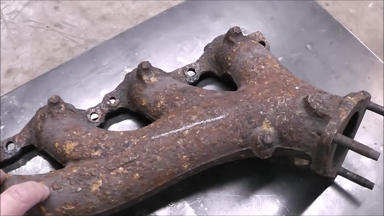 Close-up of a rusty exhaust manifold on a metal workbench, with a hand pointing to the area where a crack will be simulated for the welding demonstration.