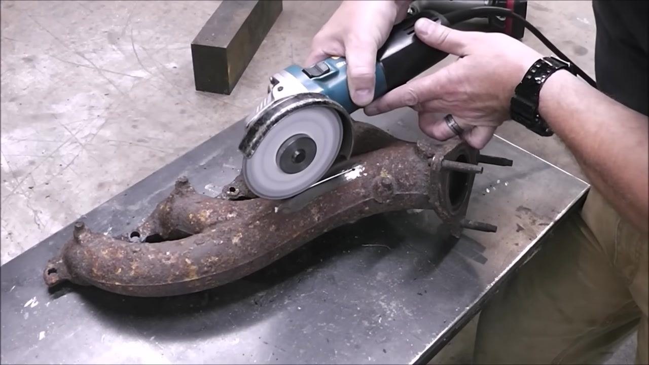 A close-up of a cast iron exhaust manifold with a shiny, ground area. Two drilled holes are visible at each end of a dark crack, and a metal pointer indicates the crack's path.