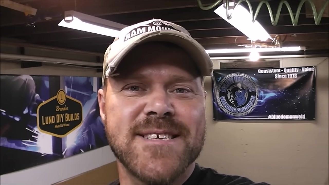 Presenter referencing a previous MIG welding video playing on a monitor in the background. Visible are patriotic star decorations, a wooden letter "B", and a cup with a straw on the desk.