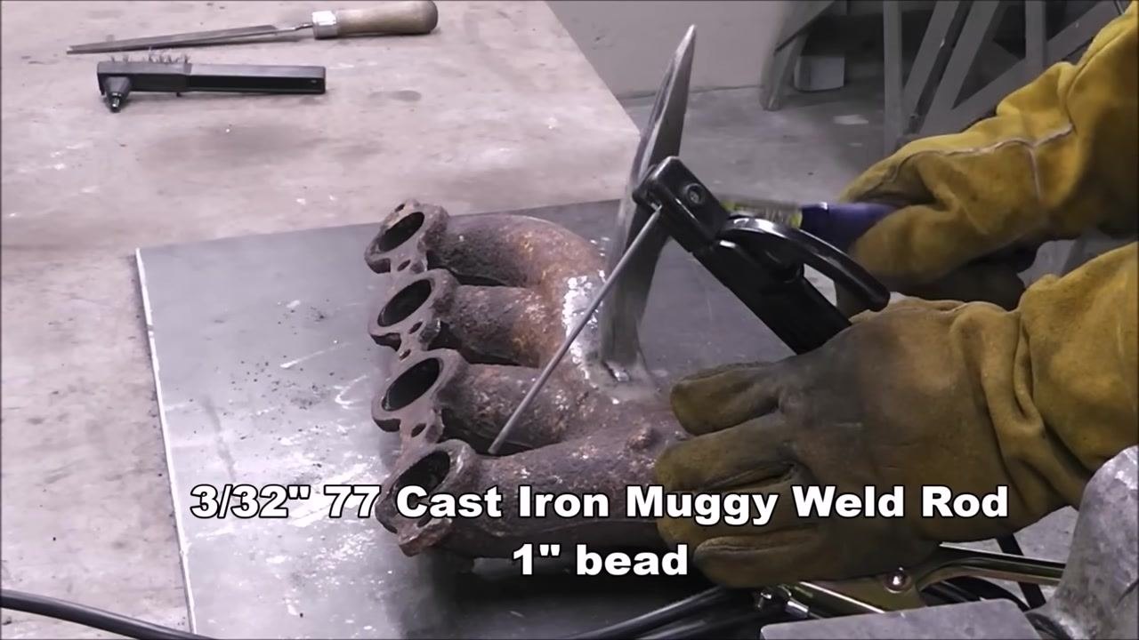 A gloved person is preparing to weld a rusty cast iron manifold using a 3/32" 77 Cast Iron Muggy Weld Rod. The rod is held in an electrode holder, and the workspace includes a file and wire brush. On-screen text: '3/32" 77 Cast Iron Muggy Weld Rod, 1" bead'