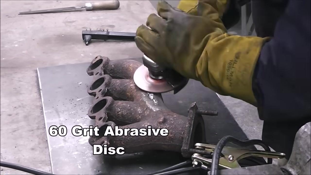 A person is using an 80 grit flap wheel on an angle grinder to finish the weld on a cast iron manifold. On-screen text: '80 Grit Flap Wheel'