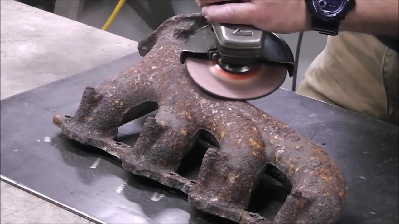 A person prepares to perform a spark test on the cleaned exhaust manifold, with a hand resting on the part and the grinder nearby. The workbench and surrounding workshop are visible.