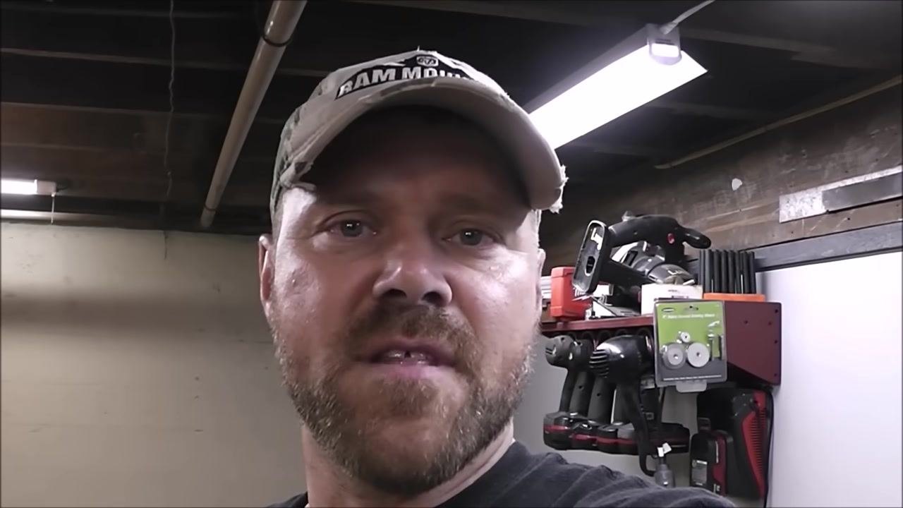 A person in a workshop, wearing a cap, is speaking directly to the camera. Behind him is a red tool rack with power tools, a green package of replacement wheels/discs, and other workshop equipment. The setting is a well-lit workshop with exposed beams.