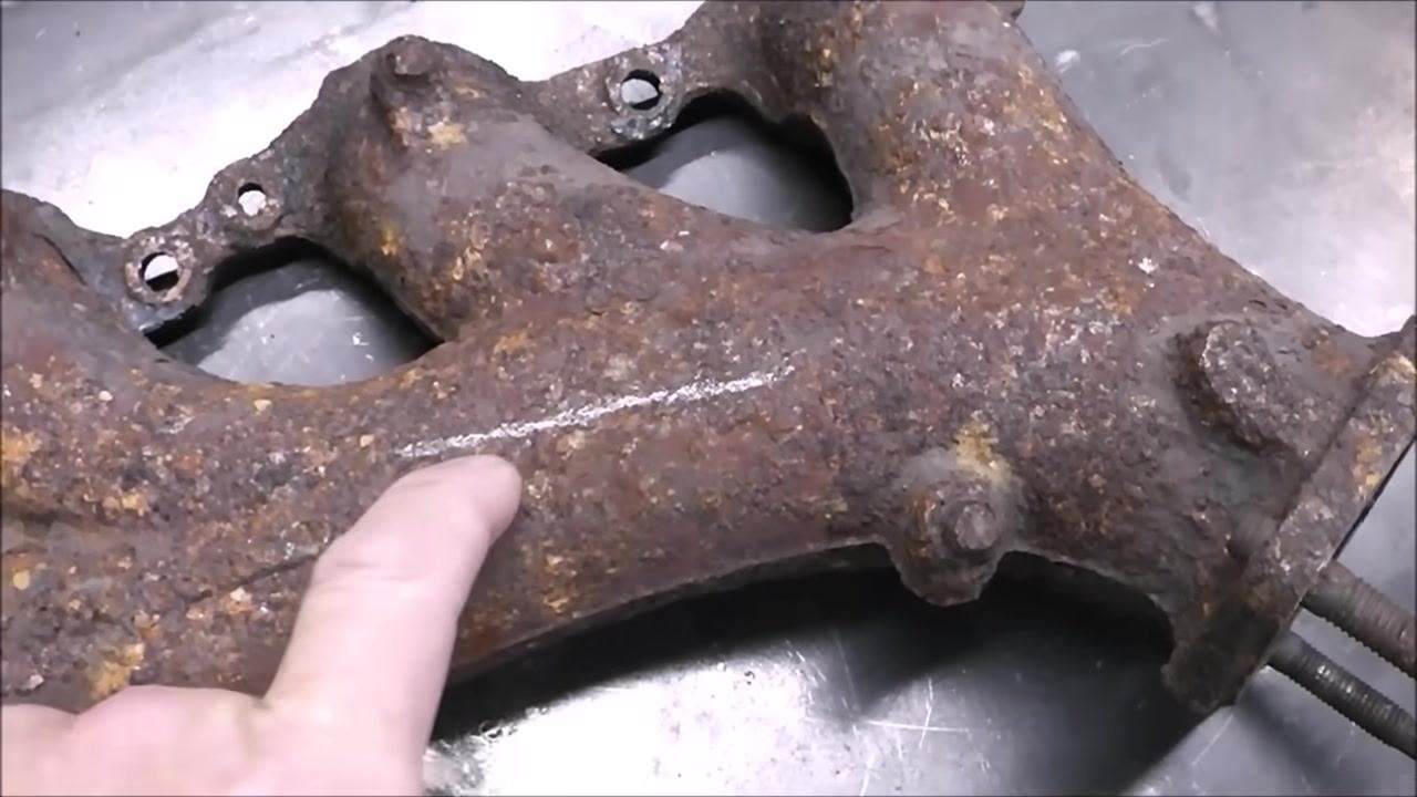 A person uses an angle grinder with a 40-grit abrasive wheel to clean a rusty exhaust manifold on a metal workbench. The grinder is in contact with the surface, removing rust.