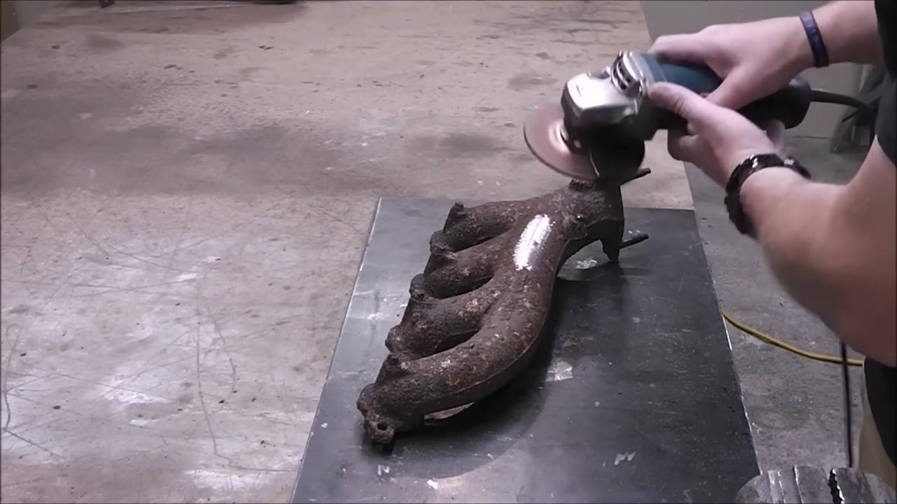 A person uses an angle grinder on the cleaned area of the exhaust manifold, producing visible sparks. The shiny, ground area contrasts with the rusty surface.