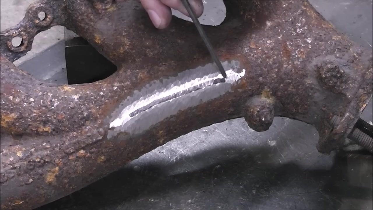 A close-up of the exhaust manifold with a shiny, ground area. A metal pointer marks the end of the crack, preparing for drilling.