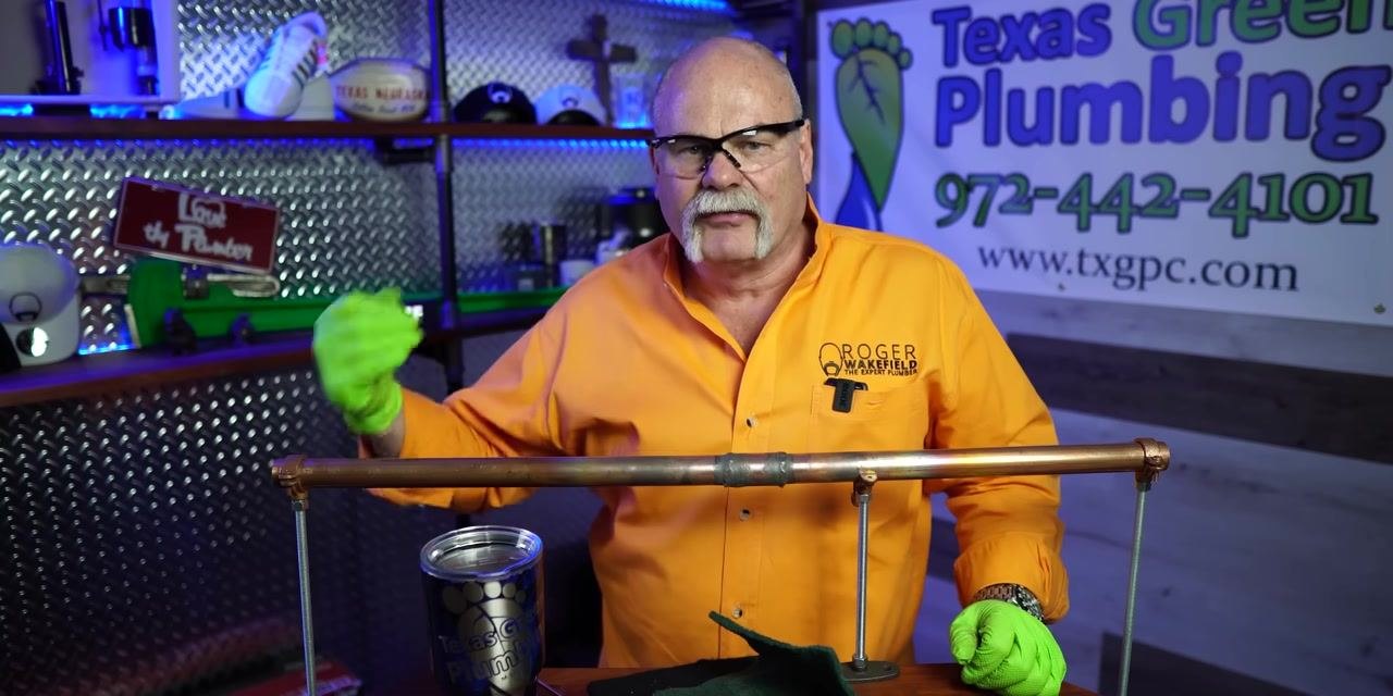Instructor in orange shirt and green gloves, gesturing with left hand while standing behind the cleaned copper pipe joint. Texas Green Plumbing banner and organized workspace visible.