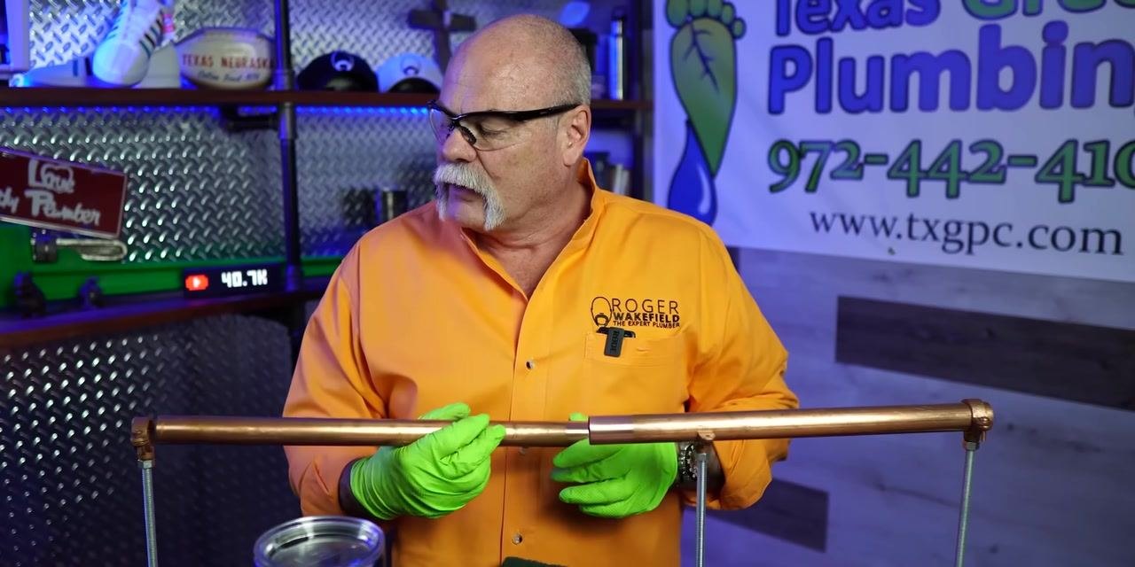 A person positions a green abrasive pad on a copper pipe in a plumbing workshop.