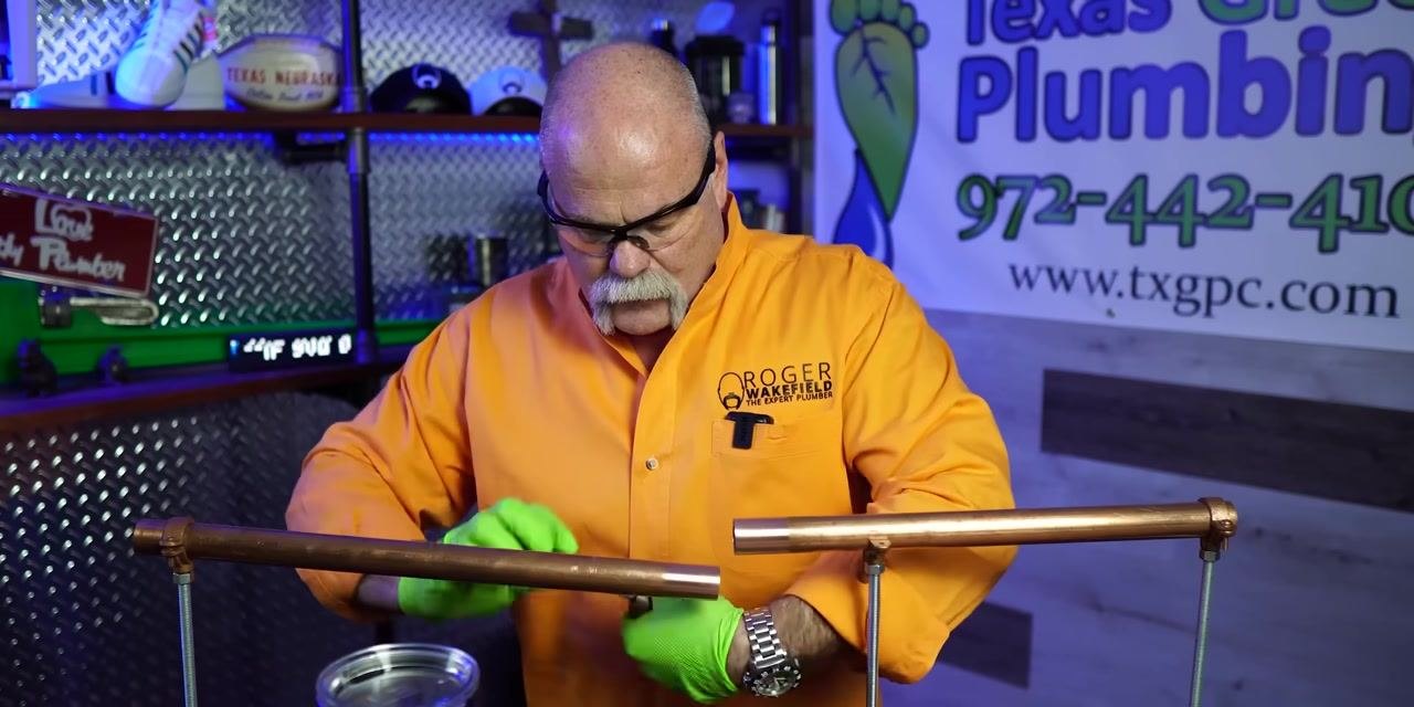 A person continues cleaning a copper pipe with a green abrasive pad in a plumbing workshop.
