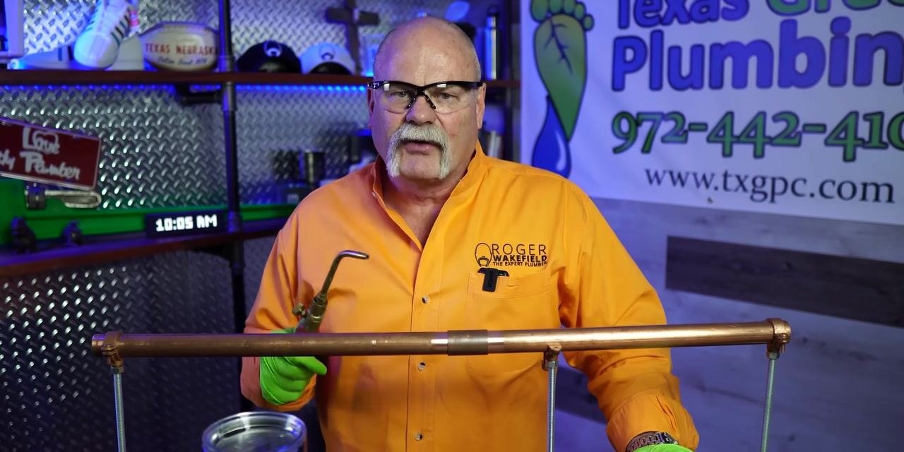 A person holds a soldering torch near a copper pipe, preparing to apply heat.
