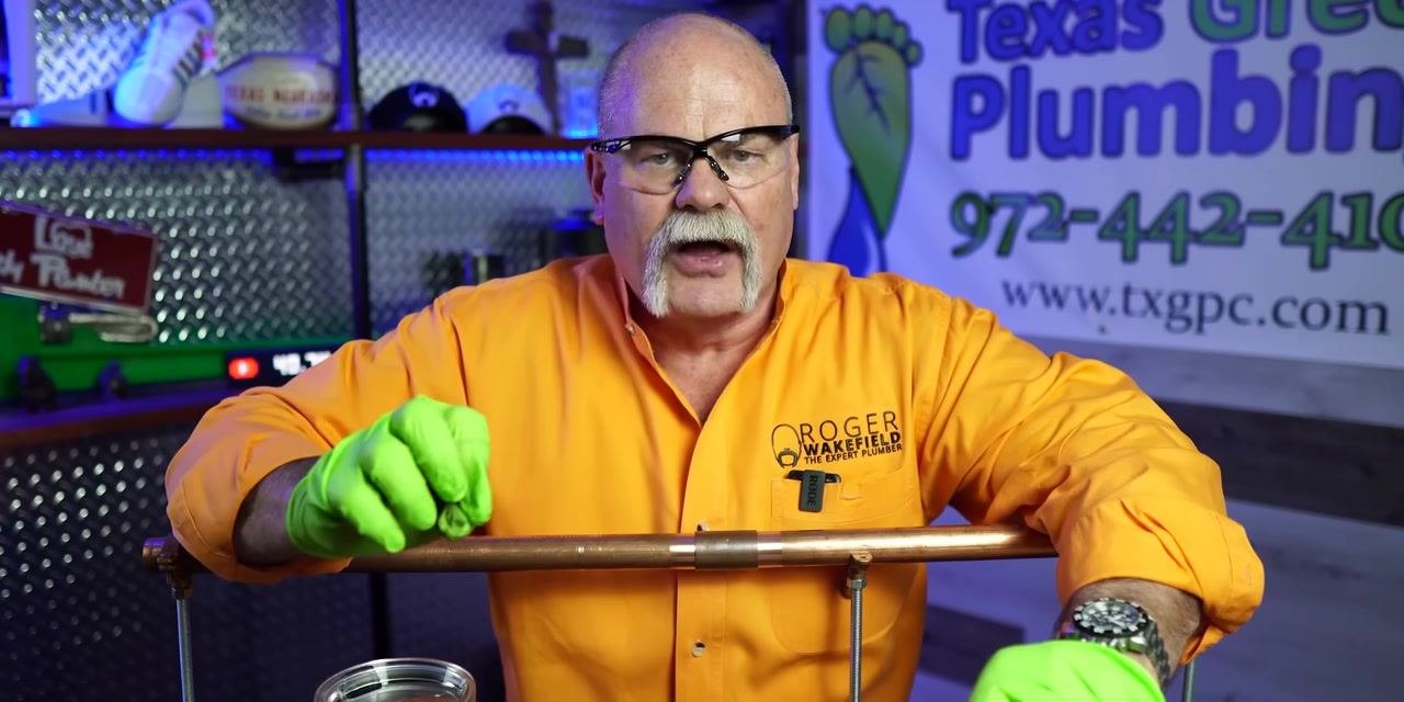 A person stands behind the workbench with both hands on a copper pipe, confirming the setup and safety equipment before soldering.