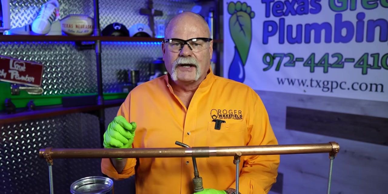 A person wearing safety glasses and green gloves stands behind a horizontally mounted copper pipe with visible joints and supports in a plumbing workshop.