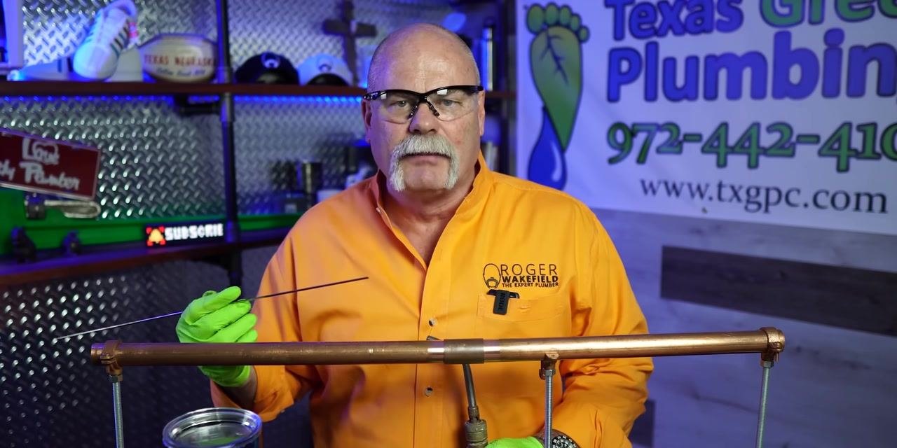 A person holds a thin solder wire near a copper pipe joint while the other hand remains near the torch.
