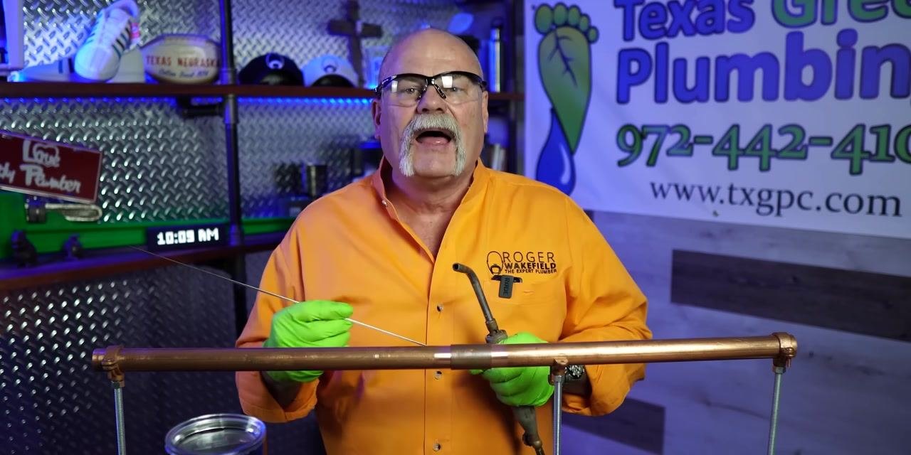 A person holds solder wire and a torch near a copper pipe joint in a plumbing workshop with branded signage and equipment.