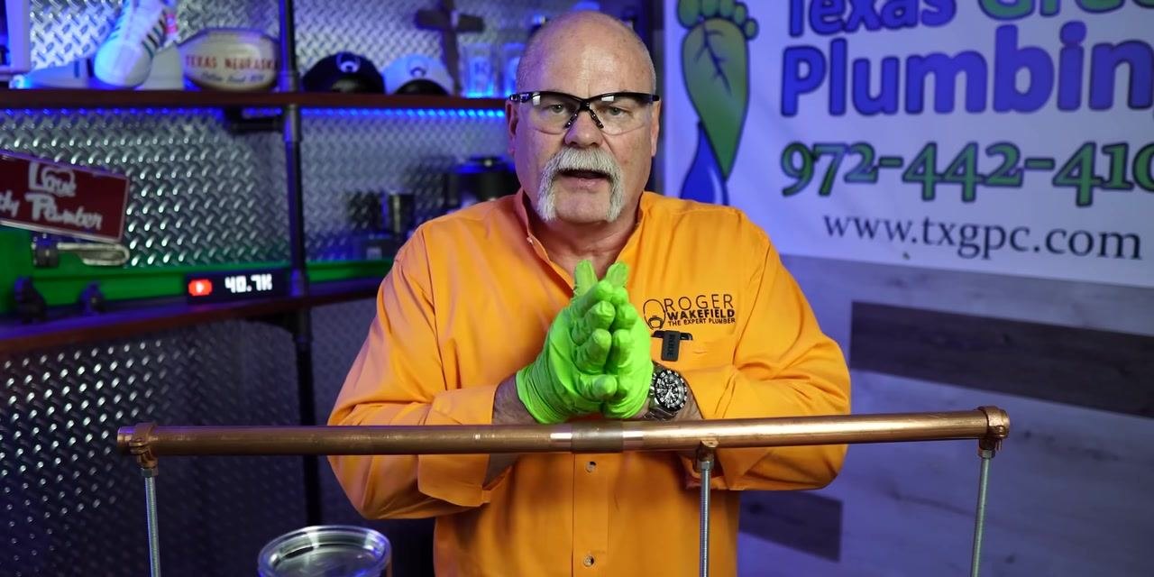 Instructor in orange shirt and green gloves holding soldering torch, preparing for soldering. Workshop background.