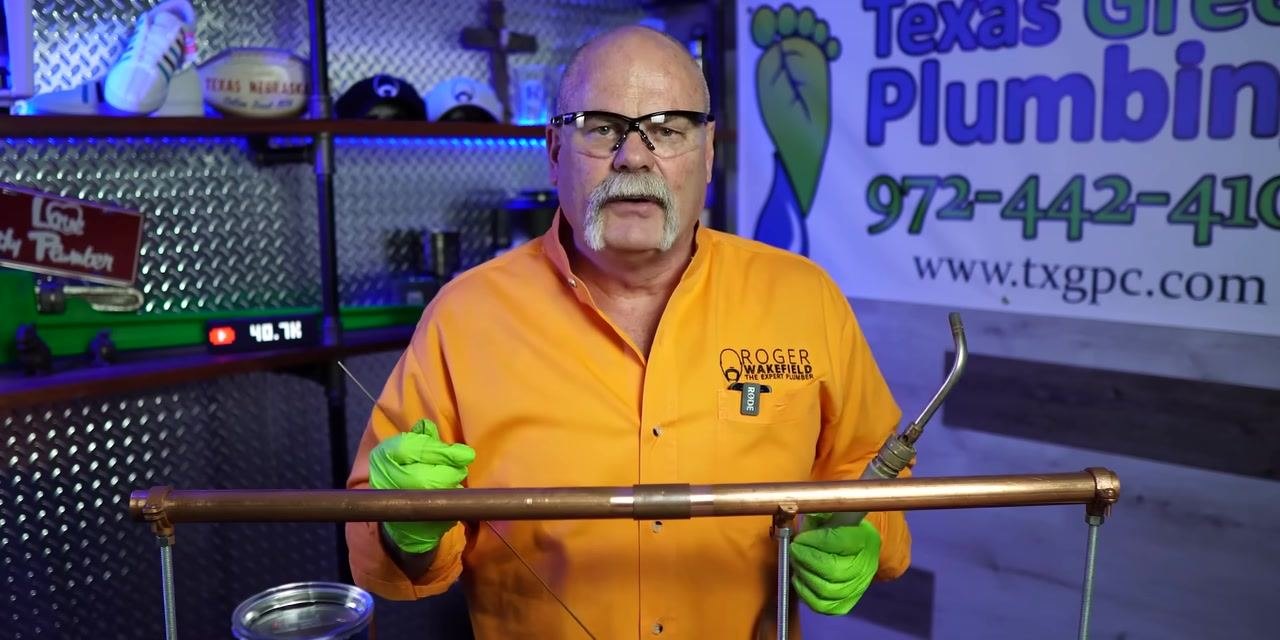 A person holds solder wire and a torch near a copper pipe joint, preparing for the next step.