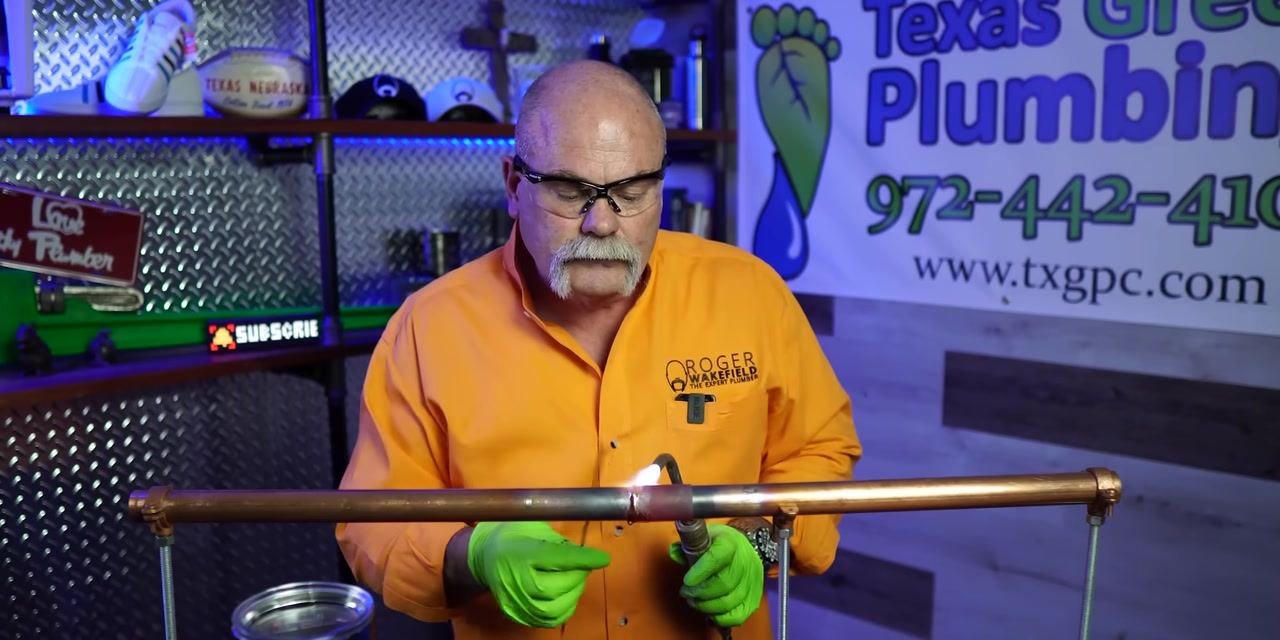 A person performs a final heating pass on a soldered copper pipe joint and inspects the seal.