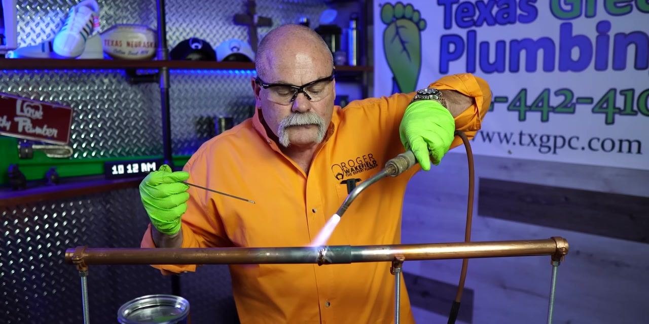 A person applies torch flame to a copper pipe joint while holding solder wire ready in a workshop with branded signage.