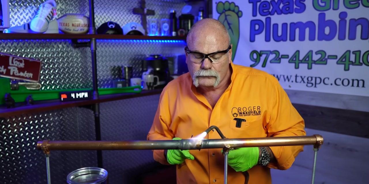A person applies solder to a heated copper pipe joint with a torch.