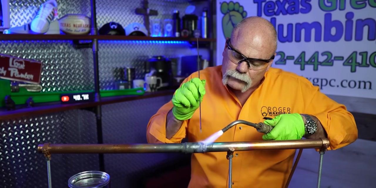 A person finishes soldering a copper pipe joint, holding a torch and solder wire.