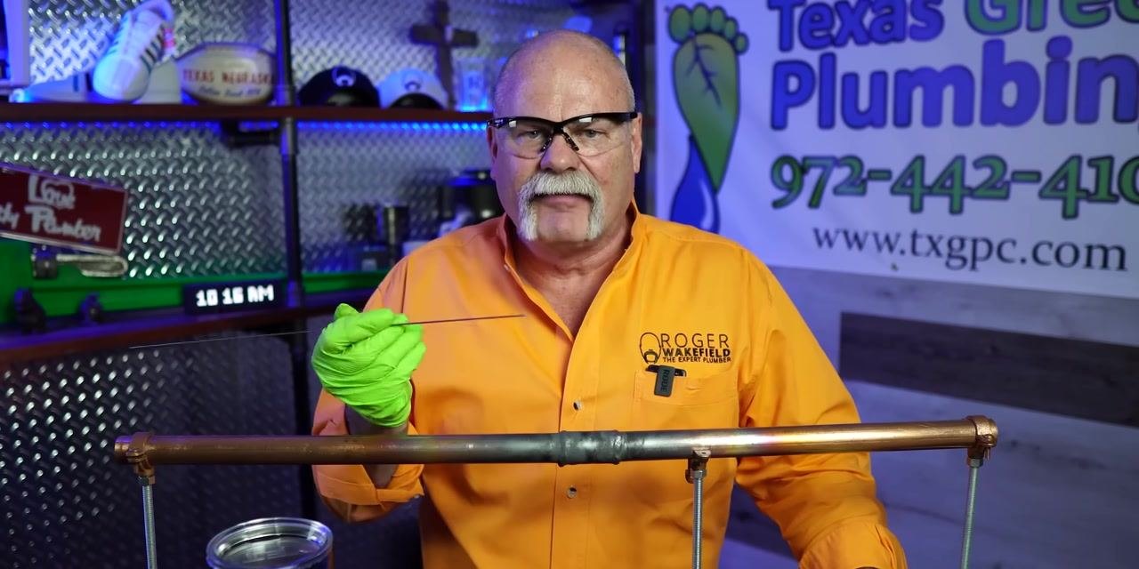 A person presents a completed soldered copper pipe joint, holding up the solder wire for reference.