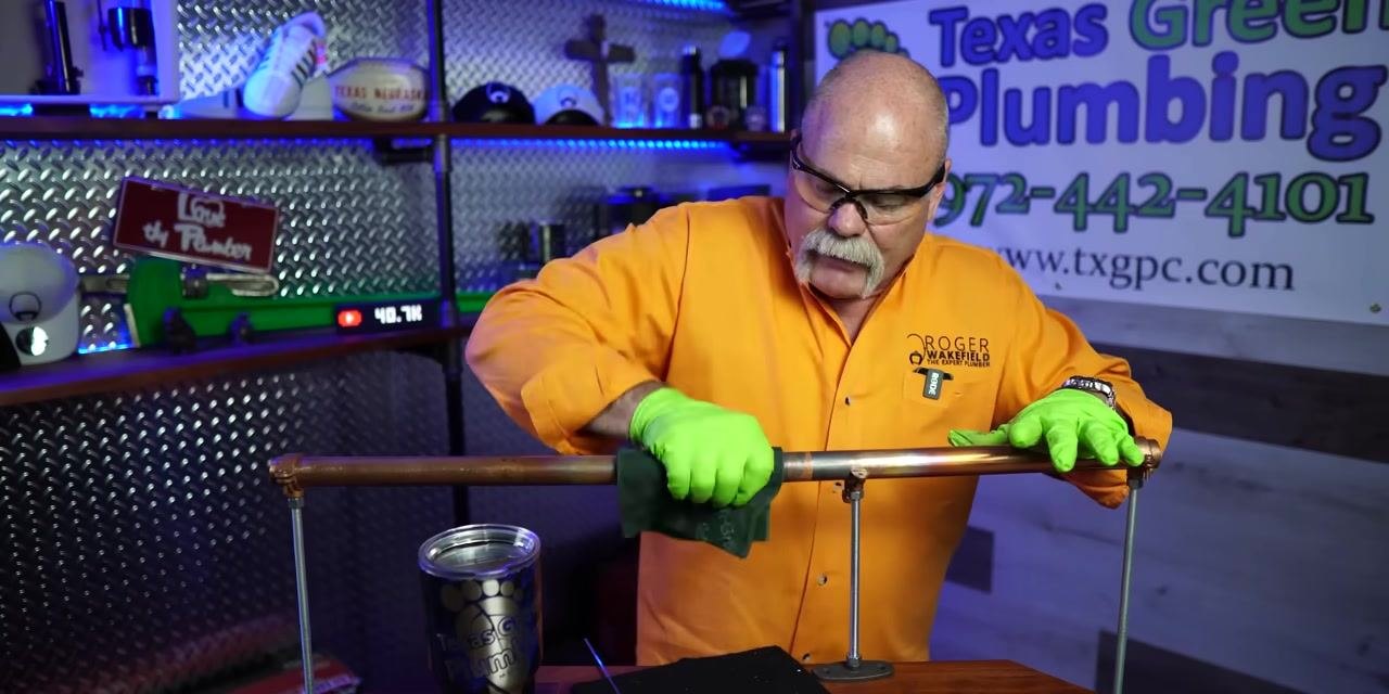 A person uses a green scouring pad to clean a soldered copper pipe joint.