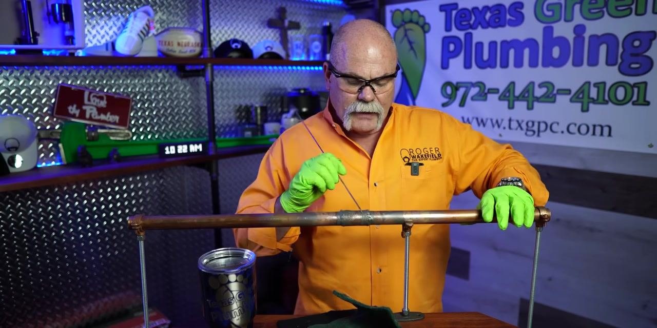 A person cleans the underside of a soldered copper pipe joint with a green scouring pad.
