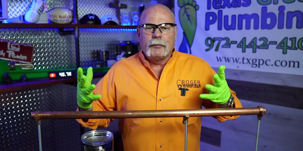 A person wearing an orange shirt and green gloves uses a pipe cleaning tool on a copper pipe, looking down at the work in a plumbing workshop.