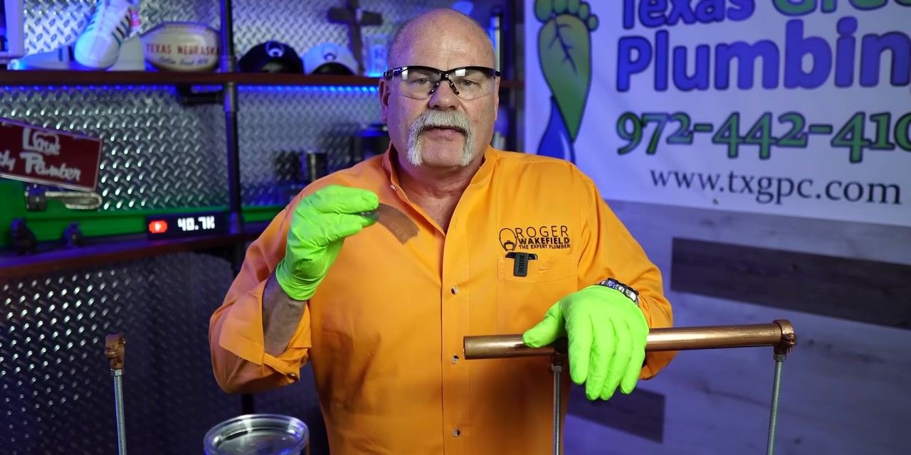 Instructor in orange shirt and green gloves holding abrasive cloth, emphasizing its use. Workshop background.