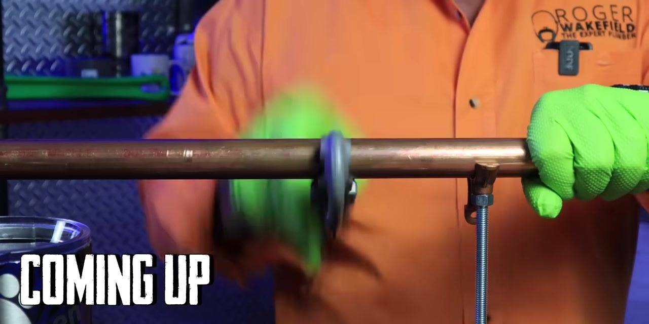 A person wearing an orange shirt and green gloves uses a pipe cleaning tool on a horizontally mounted copper pipe in a plumbing workshop with signage and tools in the background.