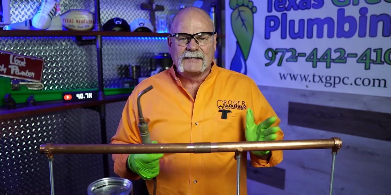 Instructor in orange shirt and green gloves explaining solder application technique, gesturing above copper pipe. Workshop background.