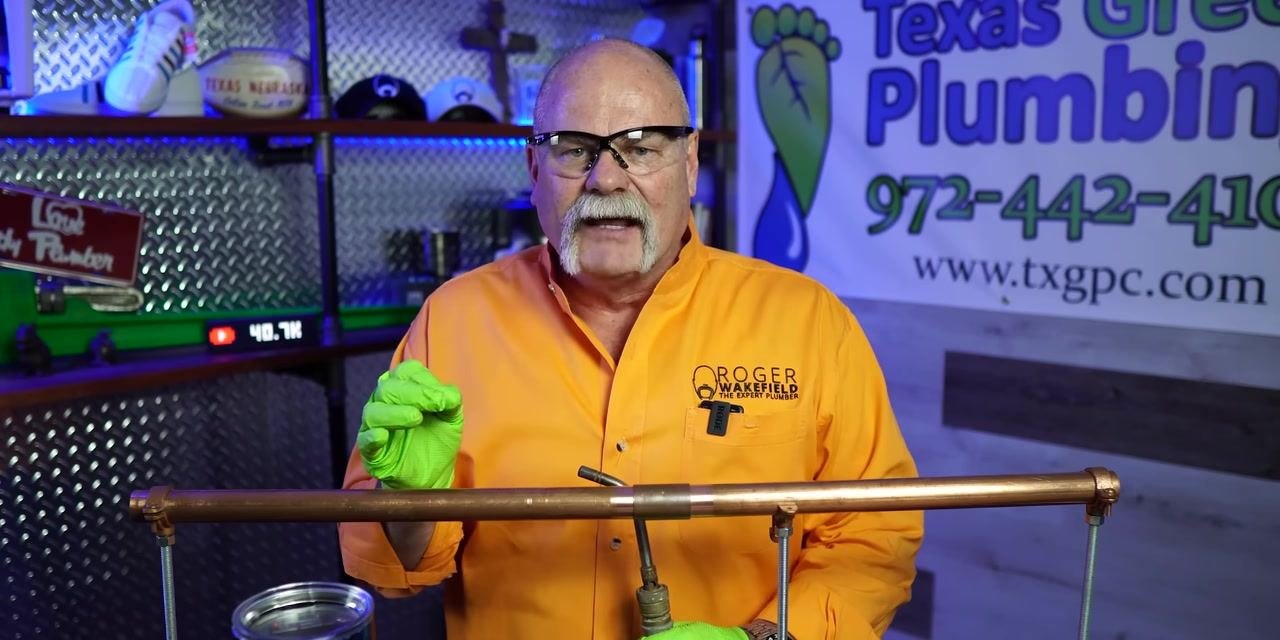The person gestures with a gloved hand while holding the torch below the pipe, emphasizing a point about the process. The workspace and setup remain consistent.