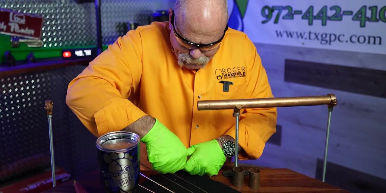 A person wearing an orange shirt and green gloves applies cleaning compound or flux to a copper fitting in a plumbing workshop.