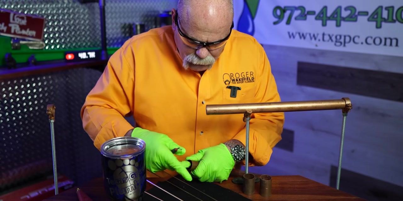A person wearing an orange shirt and green gloves attaches a copper fitting to the cleaned end of a copper pipe.