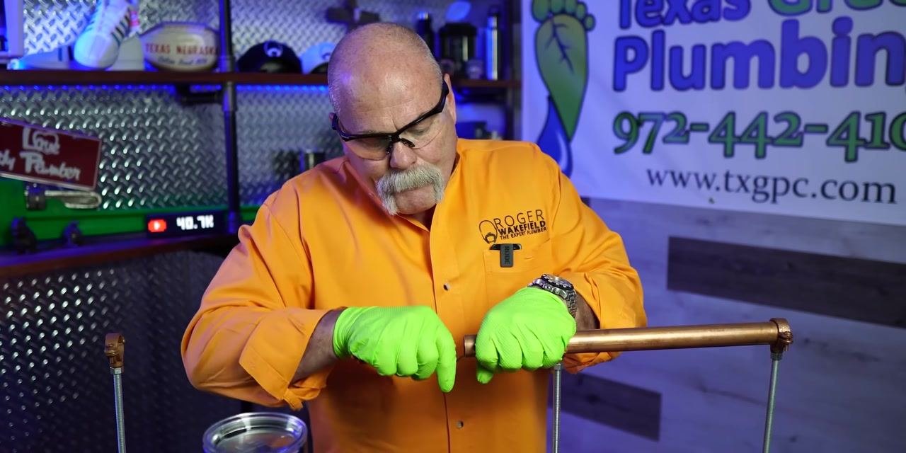 A person wearing an orange shirt and green gloves performs a final inspection and adjustment of a copper fitting on a copper pipe.