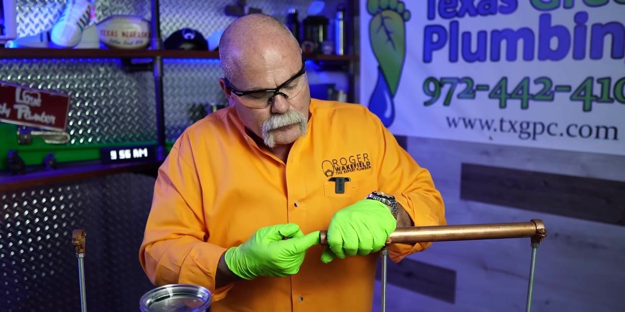 A person wearing safety glasses, green gloves, and an orange shirt uses an abrasive cloth to clean a copper pipe in a plumbing workshop.