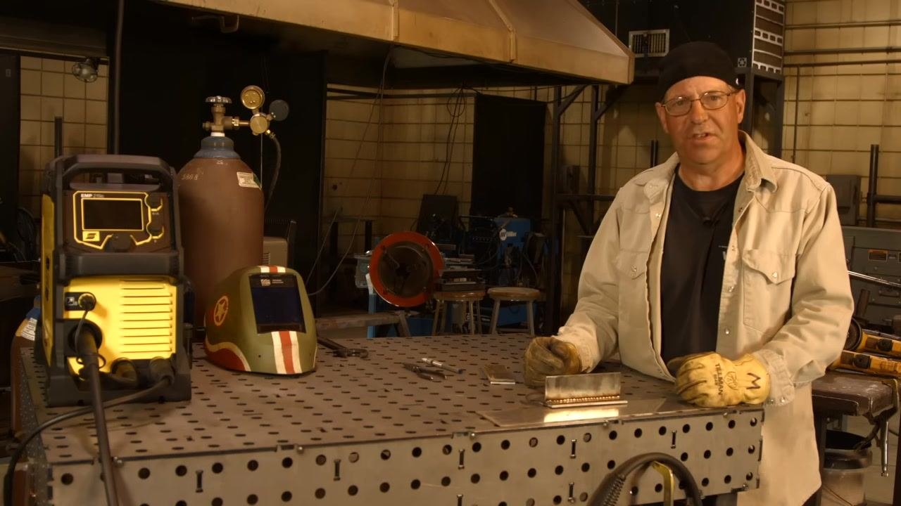 Workbench setup with welding machine, gas cylinder, helmet, tools, and completed weld sample in an industrial shop environment.