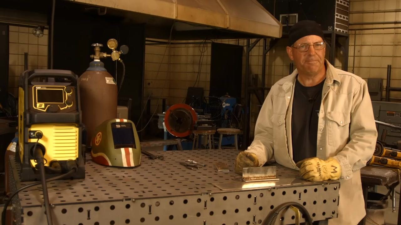 Instructor standing at welding table, smiling, with all equipment and completed weld visible, signaling the end of the session.